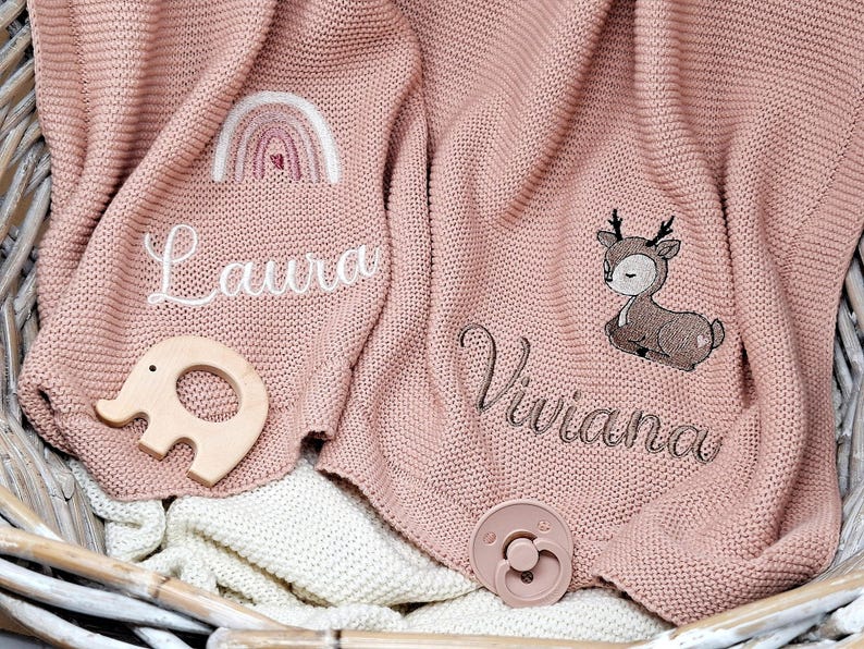 Zwei rosa gestrickte Babydecken mit gestickten Namen und Motiven. Eine Decke hat den Namen "Laura" mit einem Regenbogen, die andere "Viviana" mit einem Reh.