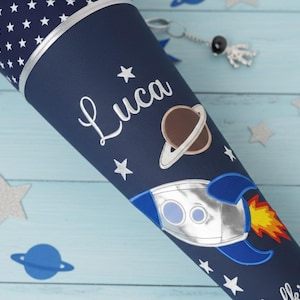 Könnte beinhalten: Eine dunkelblaue Schultüte mit weißem Sternenmuster am oberen Rand, dem Namen Luca in weißer Schreibschrift, einem braunen Planeten mit Ringen sowie einer silbernen Rakete mit blauen Flügeln und gelb-orangem Flammenausstoß.