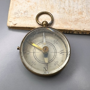 May include: A brass compass with a glass face and a silver needle. The compass is marked with the cardinal directions N, S, E, and W. The compass is on a white surface.