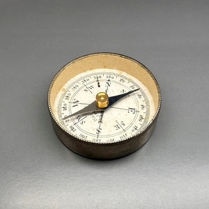 May include: A silver compass with a white face and black markings. The compass has a needle that points to north, south, east, and west. The compass is 2 inches in diameter.