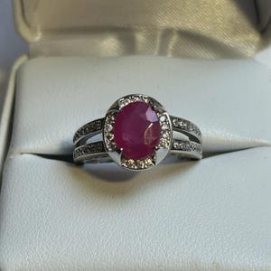 May include: A silver ring featuring a large, oval-cut ruby gemstone. The ruby is surrounded by a halo of smaller, clear gemstones. The ring has a split shank design, with additional small gemstones set into the band.