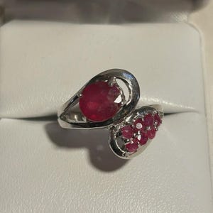 May include: A silver ring featuring a large, oval-cut ruby and a cluster of smaller rubies. The ring has a unique, asymmetrical design with a swirling band. The rubies are a deep red color, set against the bright silver.