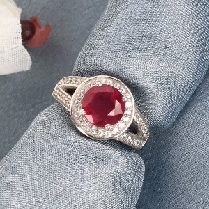 May include: A silver ring with a large, round, red ruby gemstone surrounded by smaller, white gemstones.