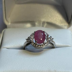 May include: A silver ring featuring a large, oval-cut ruby gemstone as the centerpiece. The ruby is a deep, rich red color. The ring is further embellished with smaller, clear gemstones around the ruby and along the band, adding sparkle and detail.