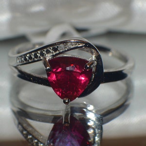 May include: A silver ring with a red gemstone and black diamonds. The ring has a unique design with the gemstone set in a triangular shape.