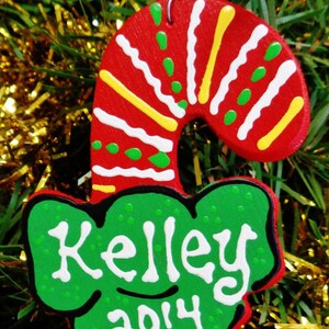 May include: A red and white candy cane Christmas ornament with green and yellow accents. The ornament has a green bow with the name "Kelley" and the year "2014" written in white.
