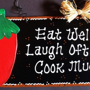 May include: A black wooden sign with white lettering that reads "Eat well, laugh often, cook much!"  A red and green painted chili pepper is on the left side of the sign.