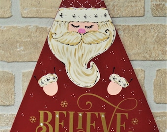 May include: A red triangular wooden sign with a white-bearded Santa Claus face, closed eyes, and a gold "BELIEVE" inscription. The sign is decorated with white snowflakes and gold glitter.