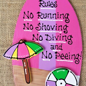 May include: Pink and purple surfboard-shaped wooden sign with the text "Pool Rules No Running No Shoving No Diving and No Peeing!"  A yellow, green, and pink umbrella and a pink, green, and purple beach ball are painted on the sign.