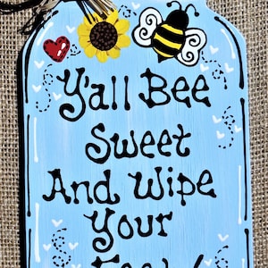 May include: A blue mason jar sign with a white bow, sunflowers, and bees. The sign reads "Y'all Bee Sweet And Wipe Your Feet!"