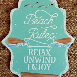May include: A turquoise blue wooden sign with white text that reads "Beach Rules: Relax Unwind Enjoy". The sign has a distressed look with white paint splatters and two brown painted driftwood pieces at the top and bottom.