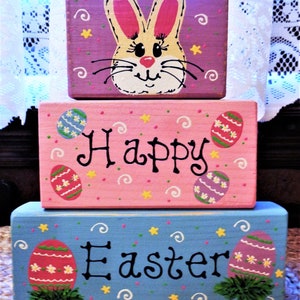 May include: A three-tiered wooden block set with a painted Easter bunny on the top tier, the middle tier says "Happy" and the bottom tier says "Easter". Each tier is decorated with painted Easter eggs and swirls.