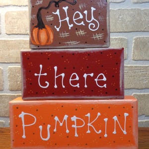 May include: Three rectangular wooden blocks stacked vertically. The top brown block features a painted orange pumpkin and the white text "Hey". The middle red block has white text "there" with black dots. The bottom orange block has white text "PUMPKIN".
