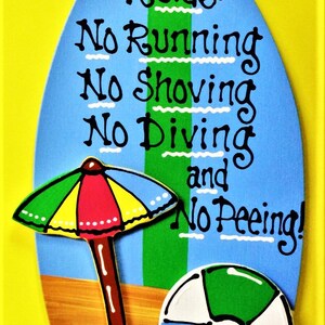May include: A blue and green surfboard-shaped sign with the text "Pool Rules: No Running No Shoving No Diving and No Peeing!"  The sign also features a beach umbrella and a beach ball.