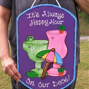 May include: Purple wooden sign with a blue border and the text "It's Always Happy Hour On Our Deck!"  The sign features two painted cocktail glasses, one green and one pink, with a lime wedge and a strawberry on top. A colorful umbrella is between the glasses.