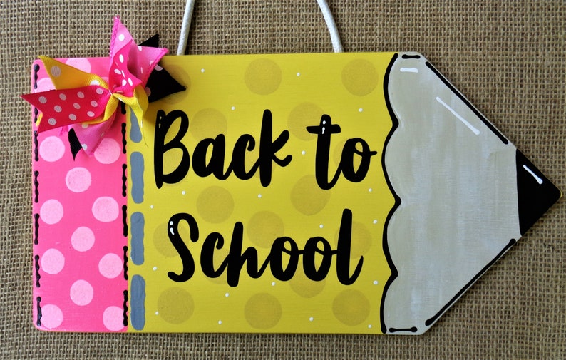 May include: A yellow pencil-shaped wooden sign with a pink and white polka dot ribbon bow. The sign says "Back to School" in black cursive lettering.