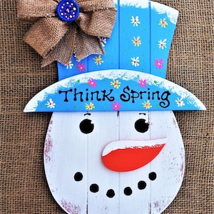 May include: A wooden snowman-shaped sign with a blue hat and a burlap bow. The sign reads "Think Spring" and features a carrot nose, black eyes, and a mouth made of dots. The hat is decorated with painted flowers.