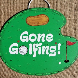 May include: Green wooden sign with a golf course design. The sign says "Gone Golfing!" with a golf ball and flag.