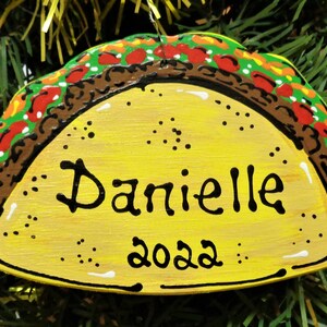 May include: A yellow painted wooden taco ornament with brown, red, and green details. The ornament is personalized with the name "Danielle" and the year "2022".