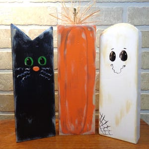 May include: Three wooden block decorations featuring a black cat with green eyes, an orange pumpkin with a raffia stem, and a white ghost with black eyes and a spiderweb detail. Each figure is painted with a hand-crafted finish and stands upright.