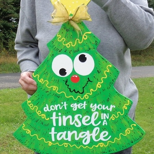 May include: A green wooden Christmas tree with a yellow star on top and a yellow bow. The tree has googly eyes and a red nose. The tree has the text "don't get your tinsel in a tangle" written on it.