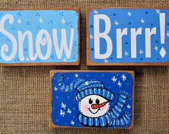 May include: Three wooden signs with a winter theme. The first sign is light blue with white snowflakes and the word "Snow" in white cursive writing. The second sign is light blue with white snowflakes and the word "Brrr!" in white. The third sign is dark blue with white snowflakes and a white snowman with a blue hat and scarf.