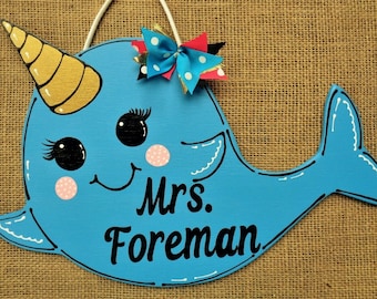 May include: A blue narwhal door hanger with a gold horn, black eyes with eyelashes, and pink cheeks. The narwhal is wearing a blue and white polka dot bow. The text "Mrs. Foreman" is written in black on the door hanger.