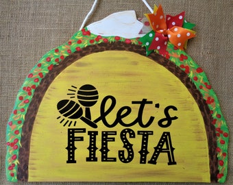 May include: A yellow wooden taco-shaped sign with a brown border and green and red accents. The sign says "Let's Fiesta" in black lettering with a maraca graphic. A red, white, and orange bow is attached to the top of the sign.