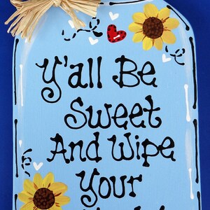 May include: A blue mason jar with a white lid and a straw bow. The jar has white hearts and sunflowers painted on it. The text on the jar reads "Y'all Be Sweet And Wipe Your Feet!"