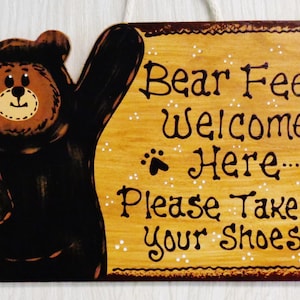 May include: A wooden sign with a brown bear illustration and the text "Bear Feet Welcome Here... Please Take Off Your Shoes!" The sign has a brown border and white polka dots.