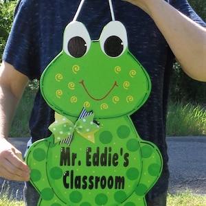 May include: Green frog-shaped wooden door hanger with white eyes, a red smile, and yellow swirls. The hanger has a white ribbon with black and white stripes and yellow polka dots. The text "Mr. Eddie's Classroom" is written in black on a white circle.