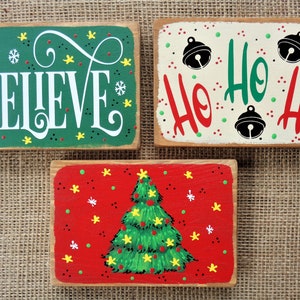 May include: Three wooden signs with Christmas themes. The first sign is green with white lettering that says "BELIEVE". The second sign is white with red and green lettering that says "Ho Ho Ho". The third sign is red with a green Christmas tree and yellow stars.