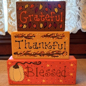 May include: Three wooden blocks stacked on top of each other. The top block is brown with orange leaves and the word "Grateful" written in orange. The middle block is orange with brown branches and the word "Thankful" written in brown. The bottom block is red with a white pumpkin and the word "Blessed" written in brown.