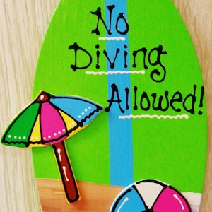 May include: A green surfboard-shaped sign with the text "No Diving Allowed!" in black. The sign has a blue stripe down the center and is decorated with a colorful beach umbrella and a beach ball.