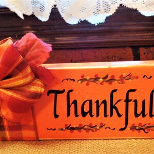 May include: Orange wooden block sign with the word "Thankful" painted in black. The sign is decorated with a red and orange plaid bow, a fall leaf, and a painted white pumpkin with a brown stem.