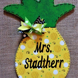 May include: A yellow pineapple-shaped wooden door hanger with a green leafy top and a white polka dot and yellow swirl pattern. The door hanger has a black bow with white polka dots and a black and white chevron bow. The text "Mrs. Stadtherr" is written in black on the front of the door hanger.