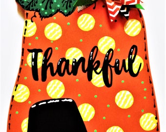 May include: Orange pumpkin-shaped door hanger with a black pilgrim hat, yellow and white polka dots, and the word "Thankful" written in black. The pumpkin has a green leaf with white polka dots and a red, white, and black bow.