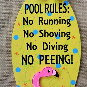 May include: A yellow oval-shaped wooden sign with a pink flamingo pool float attached to the bottom. The sign has the text "POOL RULES: No Running No Shoving No Diving NO PEEING!" in black lettering. The sign is decorated with blue and pink polka dots and a green and blue bow with white polka dots.