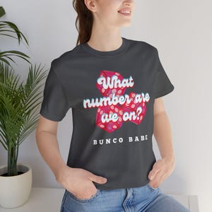 Peut inclure: Un t-shirt gris foncé avec un motif de dés et le texte "What number are we on?" et "Bunco Babe" imprimé sur le devant.