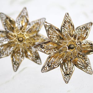 May include: Two gold-toned filigree flower-shaped earrings. Each earring features a detailed openwork design with multiple petals radiating from a central point. The earrings have a vintage aesthetic.