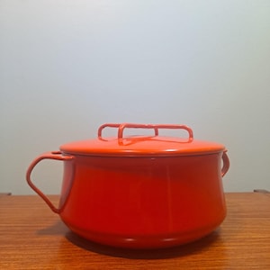 Könnte beinhalten: Ein leuchtend orangefarbener Emaille-Topf mit passendem Deckel. Der Topf hat zwei seitliche Griffe und einen Griff am Deckel. Der Topf steht auf einer Holzoberfläche.