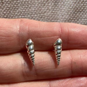 May include: A pair of silver-colored seashell stud earrings. Each earring is shaped like a spiral seashell with detailed ridges and a pointed tip. The earrings are displayed on a light brown surface.