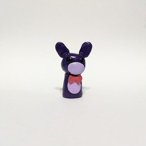 Peut inclure: Petite figurine artisanale d'un lapin violet avec de grandes oreilles et un nœud papillon rouge. Le lapin a un visage violet clair et un ventre blanc. La figurine est debout sur une surface blanche.