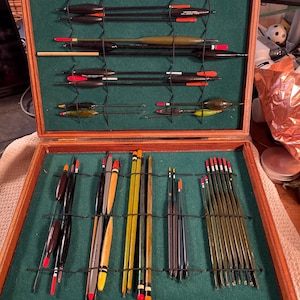 Könnte beinhalten: Eine hölzerne Angelkiste mit grünem Filzinnenraum, gefüllt mit verschiedenen Angelposen. Die Posen haben unterschiedliche Formen, Größen und Farben, darunter Rot, Gelb, Schwarz und Transparent. Die Box ist geöffnet und zeigt die organisierte Angelausrüstung.