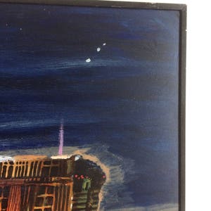 May include: A painting of a city skyline at night with a dark blue sky and a few white stars. The buildings are lit up with yellow and orange lights. The painting is framed in a black frame.