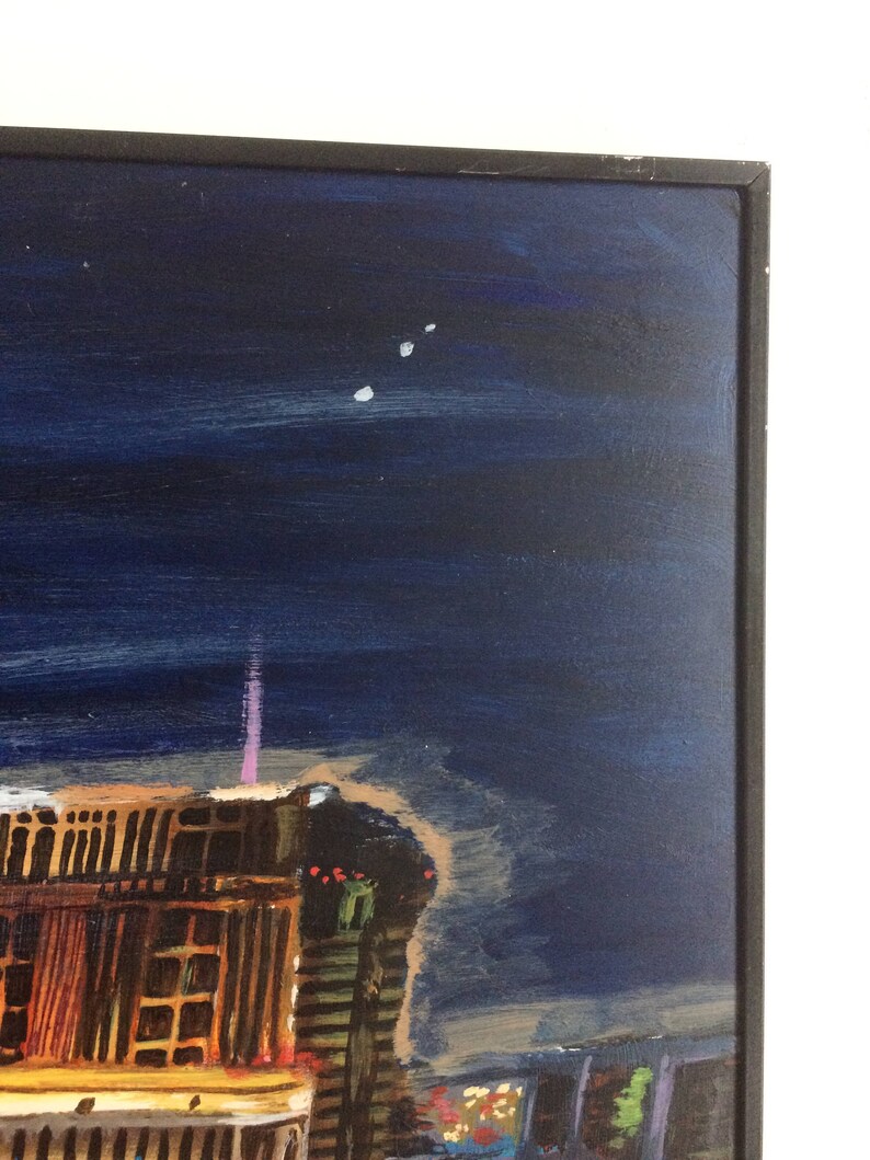 May include: A painting of a city skyline at night with a dark blue sky and a few white stars. The buildings are lit up with yellow and orange lights. The painting is framed in a black frame.