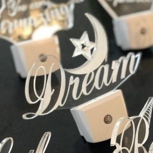 May include: A set of five white acrylic night lights with different designs. The designs include a crescent moon and star, a heart with the word "love" inside, a star with the word "amazing" inside, a crescent moon with the word "dream" inside, and a star with the word "little" inside.