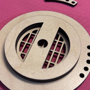 May include: Laser-cut wooden craft pieces, including a circular design with a grid pattern and a central hole, along with two curved pieces with evenly spaced holes. The pieces are light brown with black accents, set against a pink background.