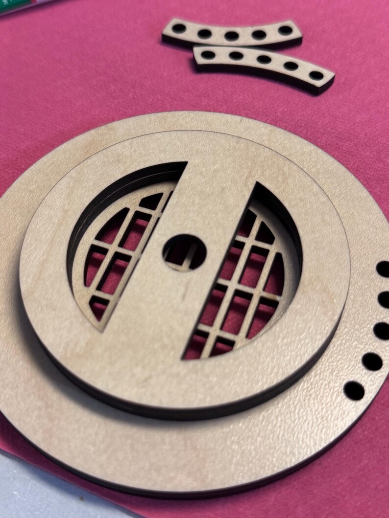 May include: Laser-cut wooden craft pieces, including a circular design with a grid pattern and a central hole, along with two curved pieces with evenly spaced holes. The pieces are light brown with black accents, set against a pink background.