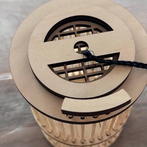 May include: A wooden lantern with a circular top featuring a decorative cutout and a black cord. The lantern's body is cylindrical, with vertical wooden supports. The design is natural and minimalist, suitable for home decor.
